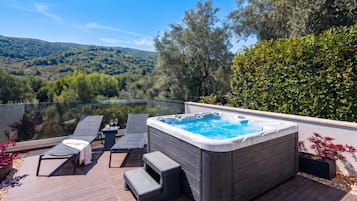 Un jacuzzi en una terraza con sillones reclinables y vistas a un lago y colinas.