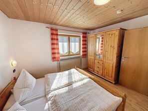 A bedroom with a wooden ceiling, a window with red curtains, a bed with striped bedding, and a wooden wardrobe. - Ferienhof Wolf by Interhome (Viechtach)