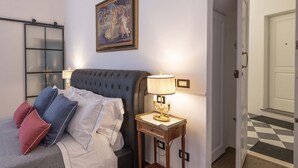 Un dormitorio con cama, almohadas, una mesita de noche con lámpara, un cuadro en la pared y un pasillo con suelo de cuadros.