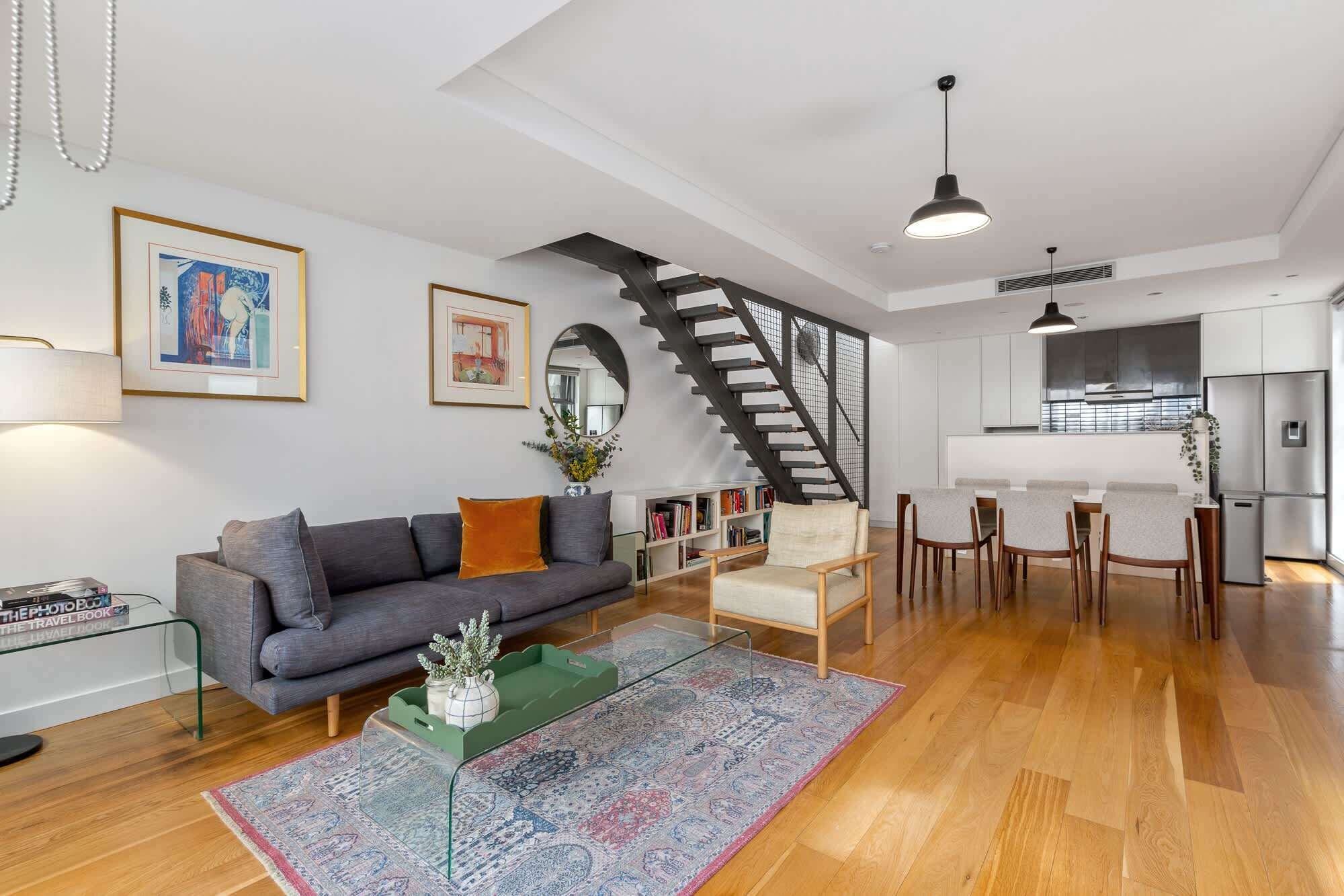 A modern living room with a gray sofa, a coffee table, a patterned rug, a staircase, and a kitchen area with a refrigerator.