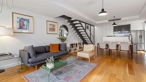 A modern living room with a gray sofa, a coffee table, a patterned rug, a staircase, and a kitchen area with a refrigerator. - Spacious 2-Bed Retreat in Rosebery (Rosebery)
