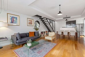 Ein modernes Wohnzimmer mit grauem Sofa, einem Couchtisch, einem gemusterten Teppich, einer Treppe und einem Küchenbereich mit einem Kühlschrank.