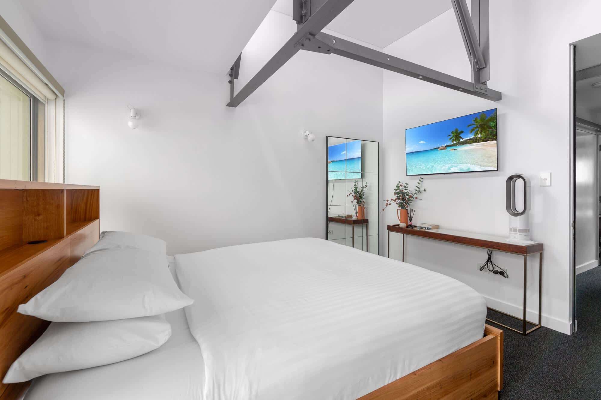 A modern hotel room with a bed, a bedside table, a mirror, and a wall-mounted TV.