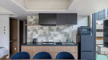 A compact kitchen with a microwave, kettle, and sink. There are two blue chairs at a wooden table, and a TV mounted above the counter.