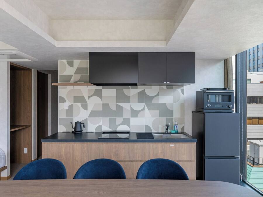 A compact kitchen with a microwave, kettle, and sink. There are two blue chairs at a wooden table, and a TV mounted above the counter.