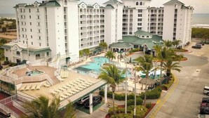 Um hotel com vários andares, área de piscina, palmeiras e vista para o oceano.