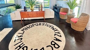 A spacious living room with a large window, a ceiling fan, a round rug with text, and wicker furniture.