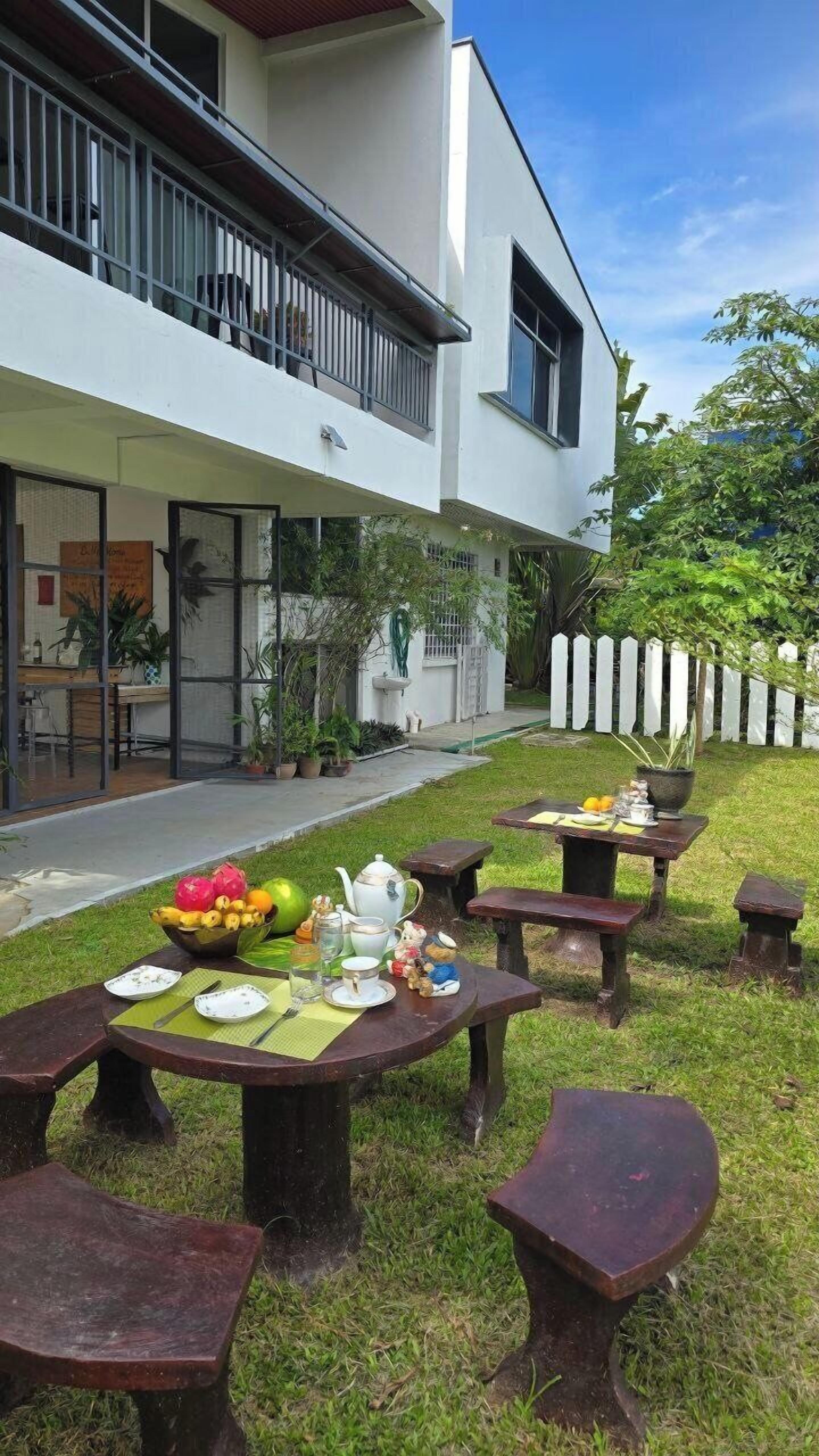 A modern building with a balcony, outdoor seating area with tables and benches, and a garden with potted plants.