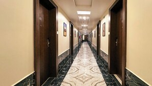 Um corredor comprido e estreito, com piso de mármore, portas de madeira e quadros nas paredes.