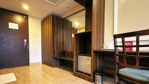 Ein Hotelzimmer mit einem Holzschrank, einem kleinen Kühlschrank, einer Metallmülltonne und einem Holzsessel.