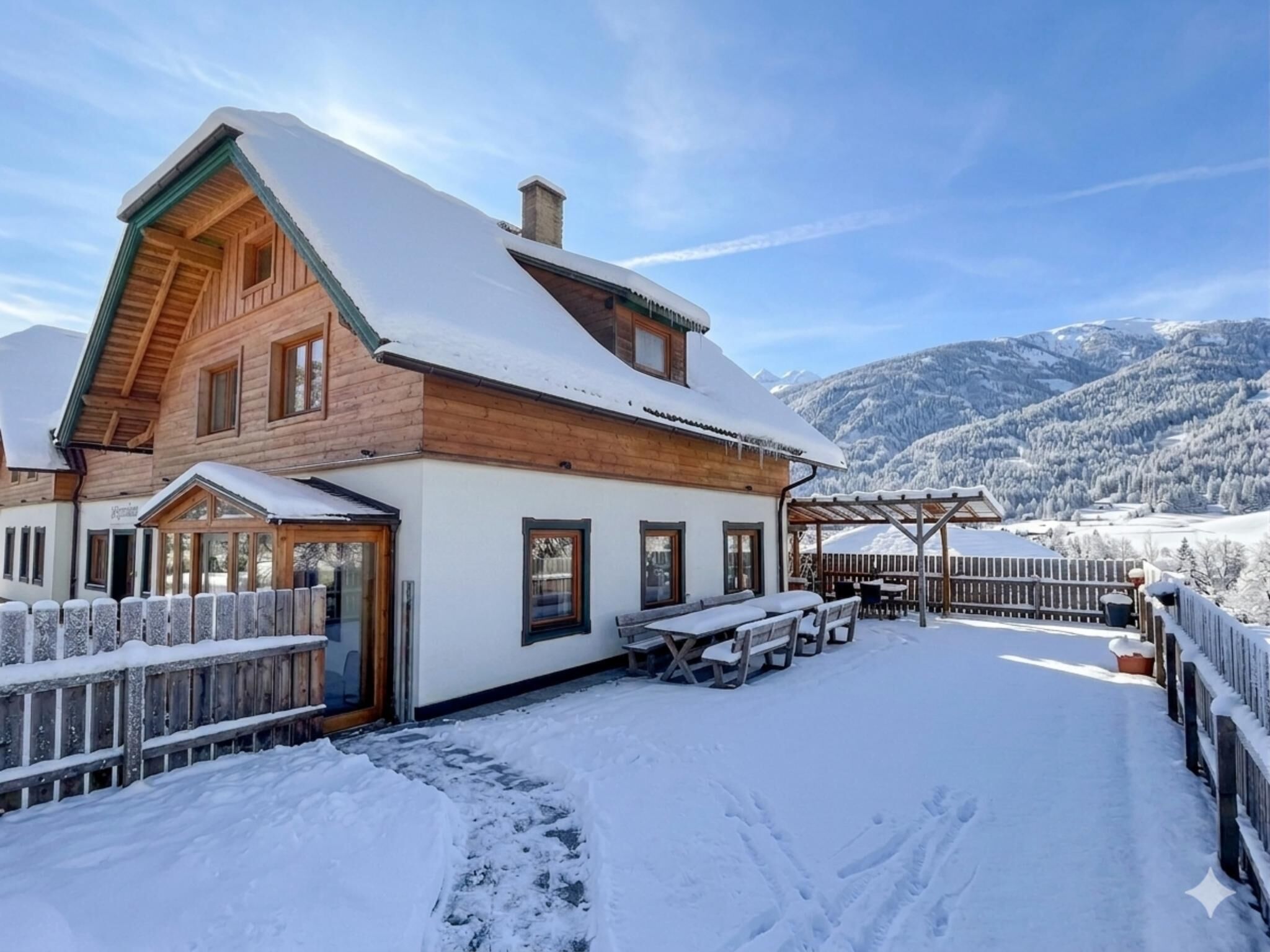 A wooden chalet with a snow-covered roof, a patio with outdoor seating, and a view of snow-covered mountains.