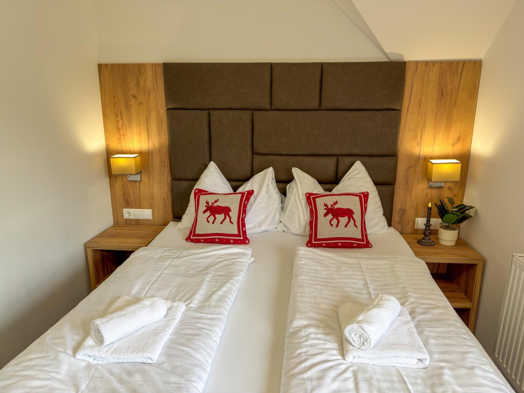 Una cama doble con sábanas blancas y dos almohadones decorativos con motivos de renos, flanqueada por cabeceros de madera y lámparas de noche.