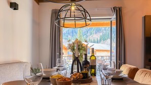 Mesa de jantar posta com taças de vinho, tigelas e tábua de queijos, com vista para uma floresta e um edifício através de uma janela.