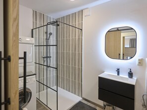 Een moderne badkamer met een douchecabine van glas, een ingebouwde wasmachine, een wastafelmeubel met spiegel en een klein wastafel.
