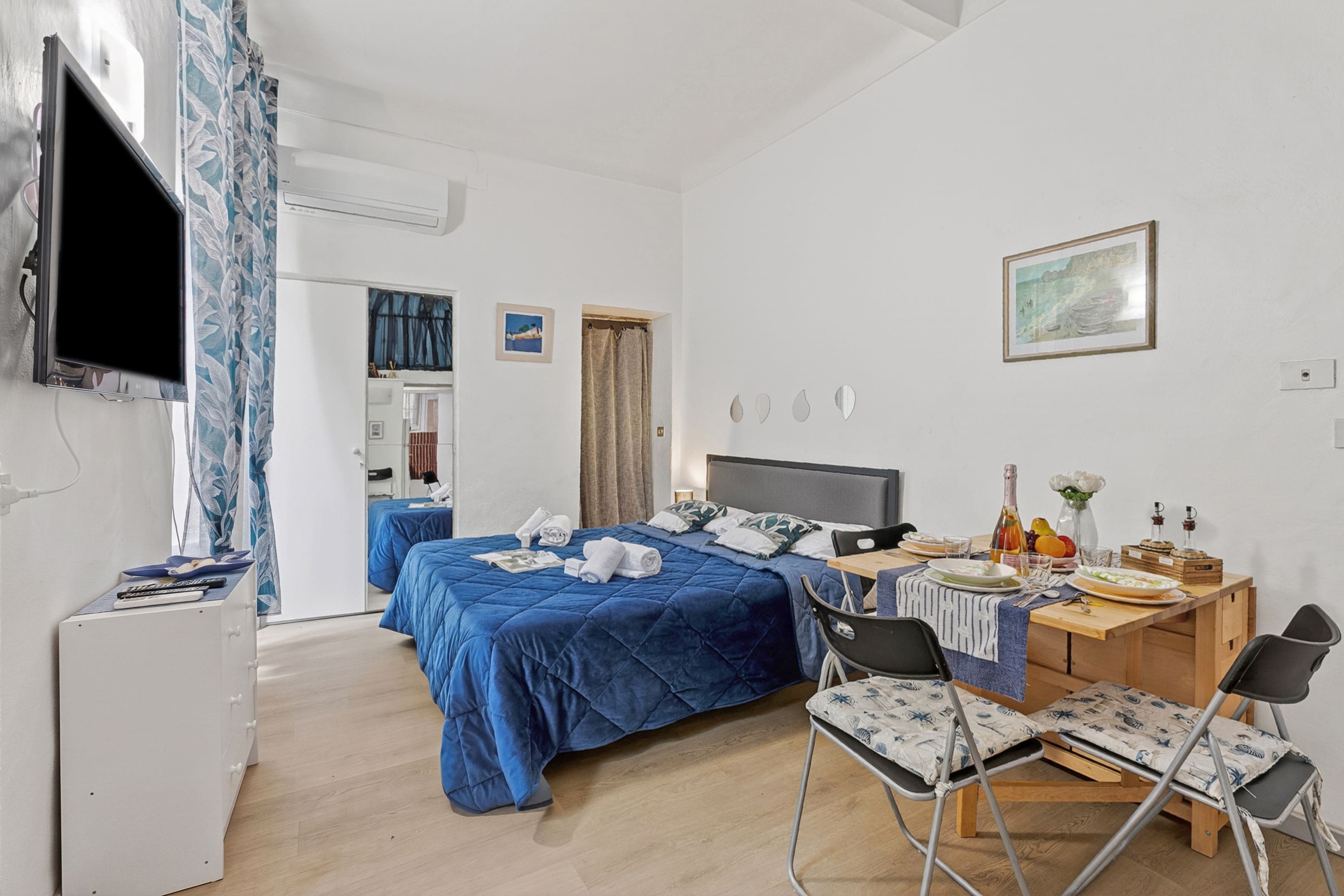 Quarto de hotel com cama, escrivaninha, cadeiras, televisão e mesa de jantar.