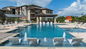 A swimming pool with multiple water jets, surrounded by lounge chairs and umbrellas, with a building in the background. - Blueground | Scofield Farms, pool, nr Walnut Creek Park (Austin)