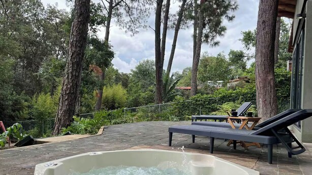 A hot tub surrounded by trees, a lounge chair, and a wooden table with a cup.