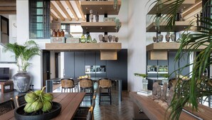 A modern kitchen with wooden shelves, a dining area with a large table and chairs, and a view of the city. - Villa Sierra (Koh Samui)