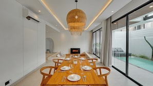 A modern dining room with a wooden table set for six, a large window with a view of a swimming pool, and a hanging light fixture. - Villa RHYS - Canggu (Canggu)