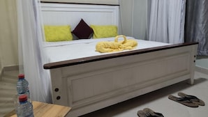 Ein Bett mit weißem Gestell, einem hölzernen Kopfteil und einem gelben Handtuch. Auf einem kleinen Tisch neben dem Bett stehen zwei Wasserflaschen.