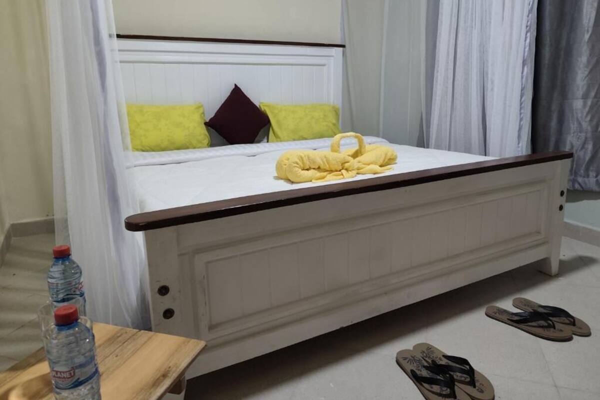 A bed with a white frame, a wooden headboard, and a yellow towel on the bed. There are two bottles of water on a small table beside the bed.