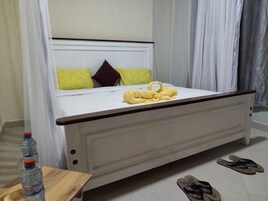 A bed with a white frame, a wooden headboard, and a yellow towel on the bed. There are two bottles of water on a small table beside the bed.