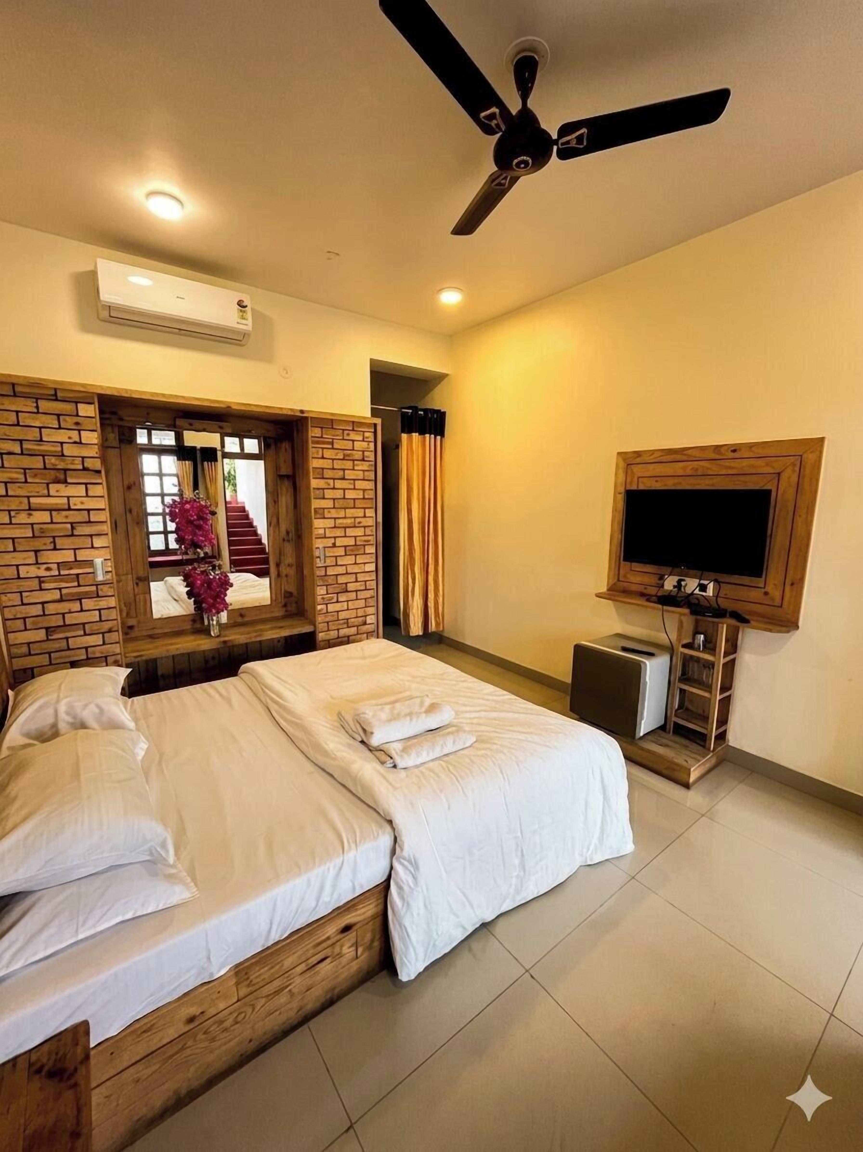 Habitación de hotel con cama, ventilador de techo, televisor y pared de ladrillo.