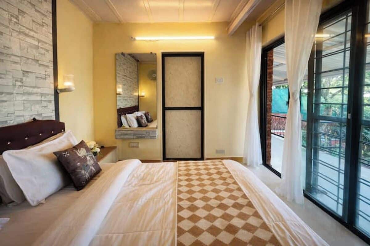 Une chambre d’hôtel avec une couvre-lit à carreaux, une grande fenêtre avec des rideaux, un miroir et un luminaire fixé au mur.
