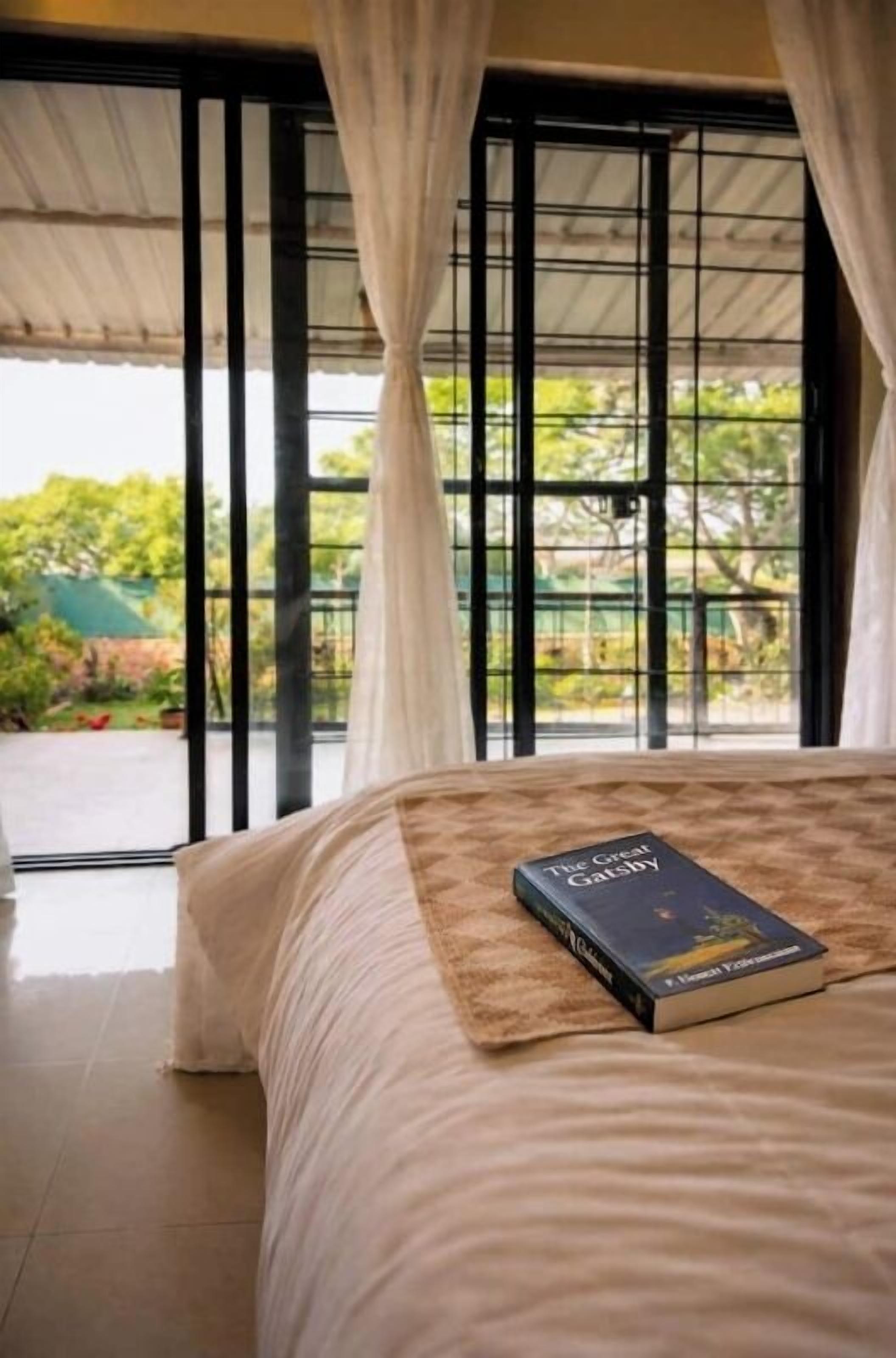 A bedroom with a bed, a book titled 'The Great Gatsby,' and a view of greenery through a window with curtains.
