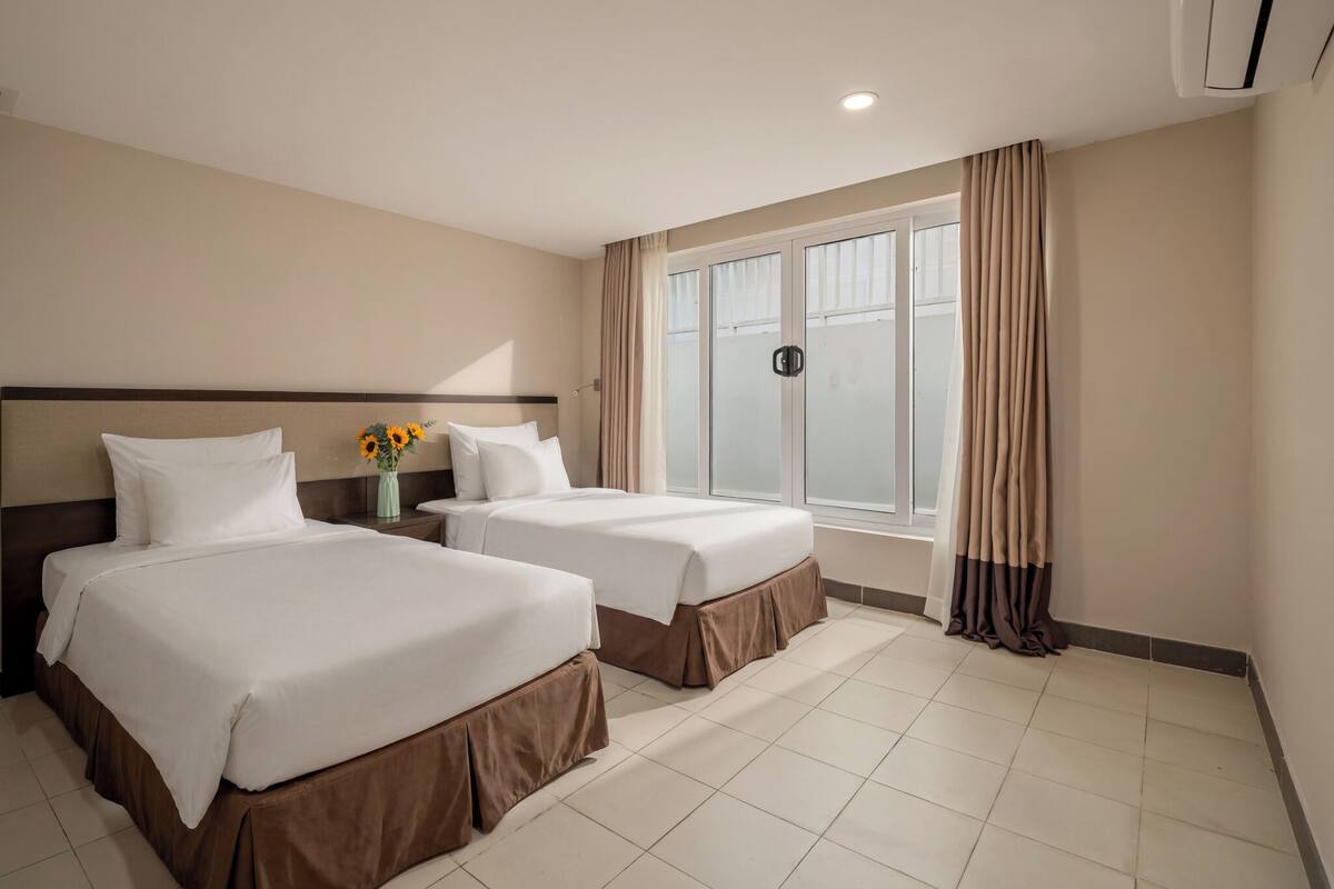 Habitación de hotel con dos camas, una mesita de noche, un jarrón con flores, una ventana con cortinas e iluminación en el techo.