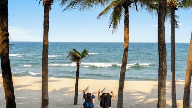 Two people with conical hats on a beach with palm trees and ocean waves. - Pool Villas Resort (Da Nang)
