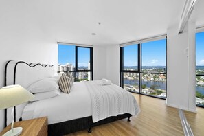 A modern bedroom with a large bed, a nightstand, a lamp, and a city view through large windows. - Panorama by Khove (Surfers Paradise)