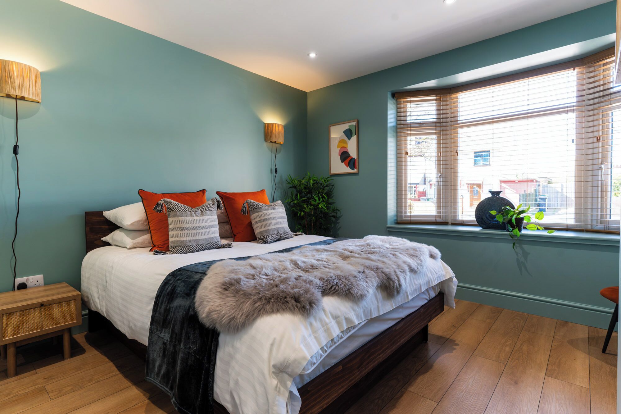 A bedroom with a bed, bedside lamp, window with blinds, and a view of the outside.