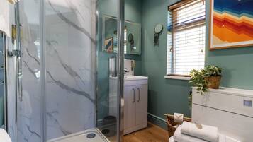 A bathroom with a glass shower enclosure, a sink with a mirror, a window with blinds, and a potted plant.