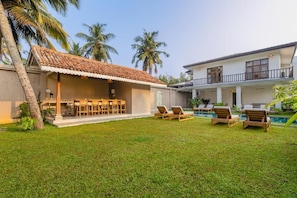A villa with a covered outdoor dining area, a pool with lounge chairs, and a two-story building with balconies.