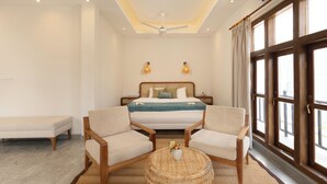 Un dormitorio con una cama, dos sillones, una mesita redonda y un ventanal con cortinas.