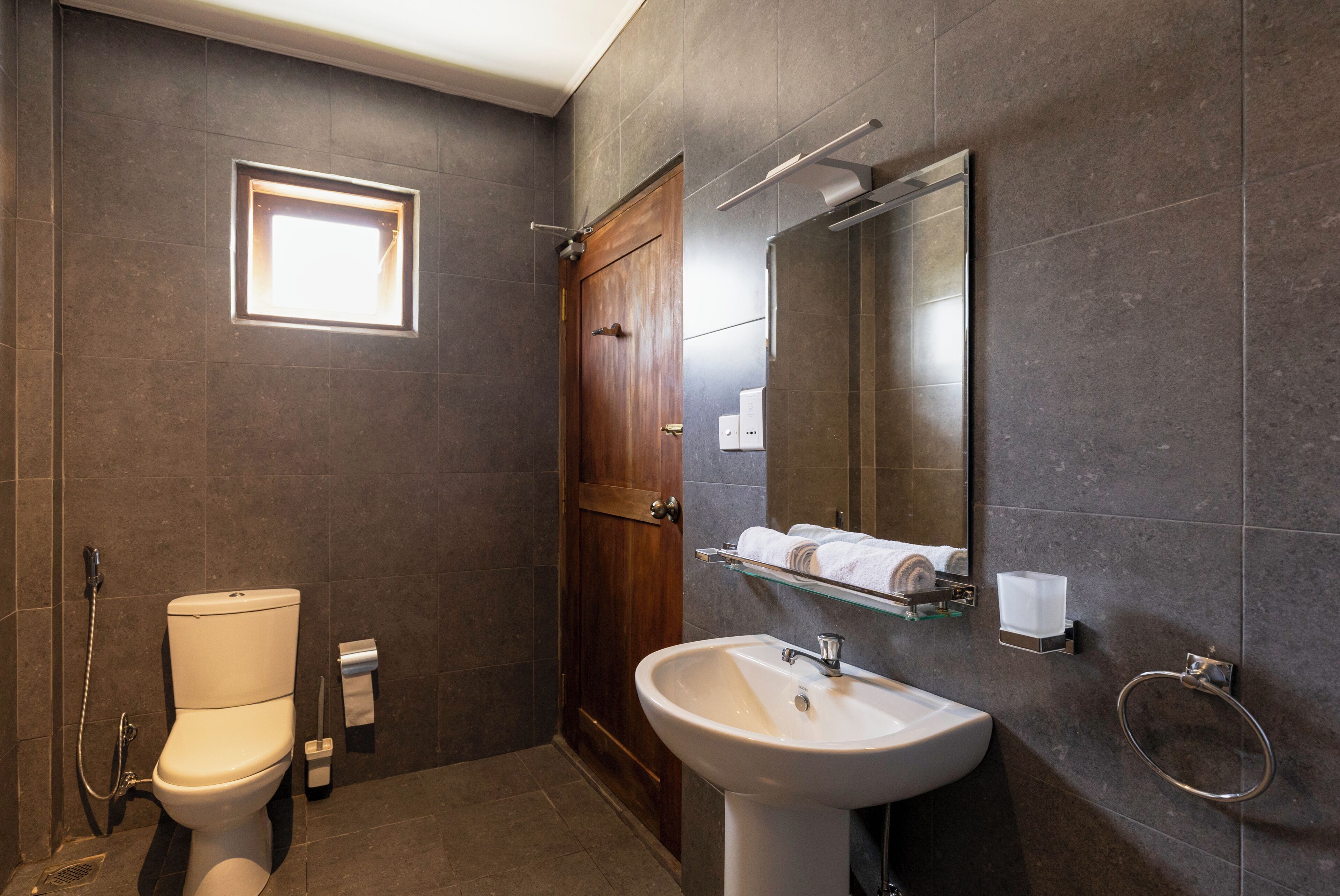 Un baño con inodoro, lavamanos, espejo, toallero y una ventana pequeña.