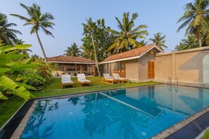 A swimming pool with lounge chairs, surrounded by palm trees and a building with a tiled roof.