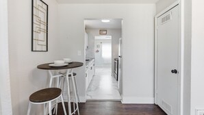 Un corridoio stretto che conduce a una cucina con mobili bianchi, una zona pranzo con sgabelli e un quadro appeso al muro.