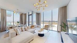 Un salón amplio con un sofá grande, una mesa de centro y una televisión. Se ven ventanas grandes con vistas a la ciudad.