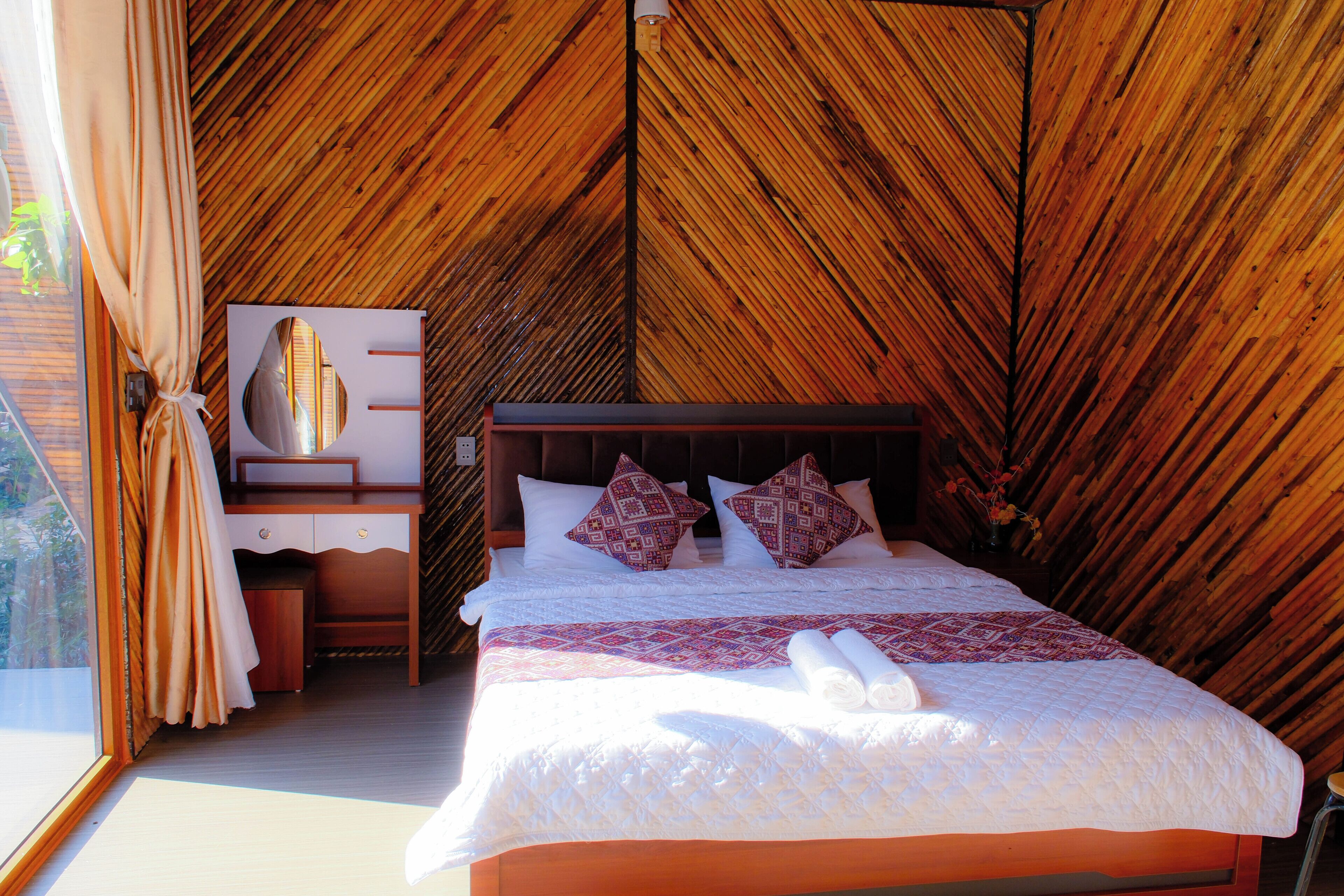 Ein Schlafzimmer mit holzgetäfelter Decke, einem Bett mit weißer Bettwäsche und gemusterten Kissen, einem Nachttisch, einem Spiegel und einem Fenster mit Vorhängen.