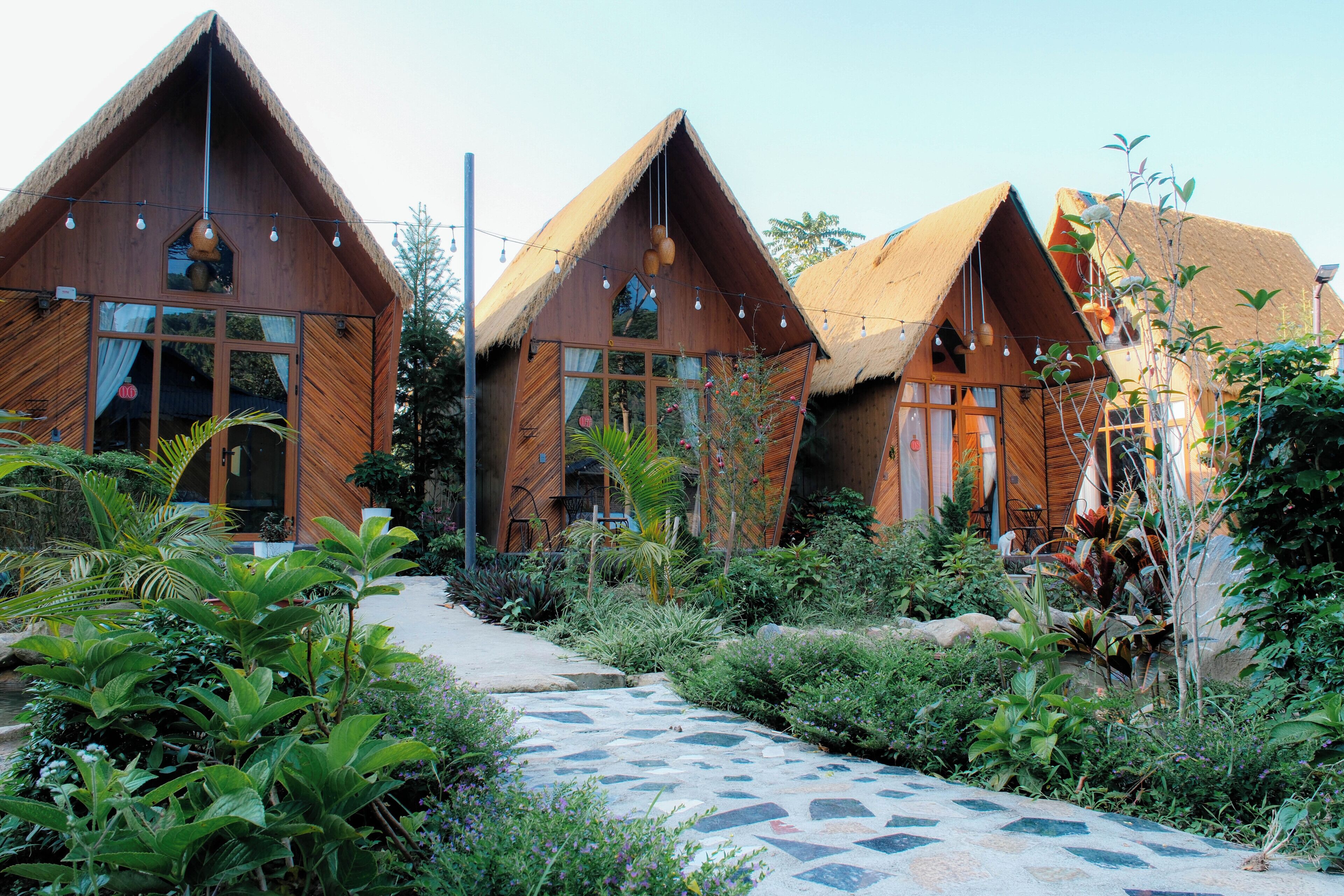 A row of wooden cabins with thatched roofs, surrounded by lush greenery and a stone pathway.