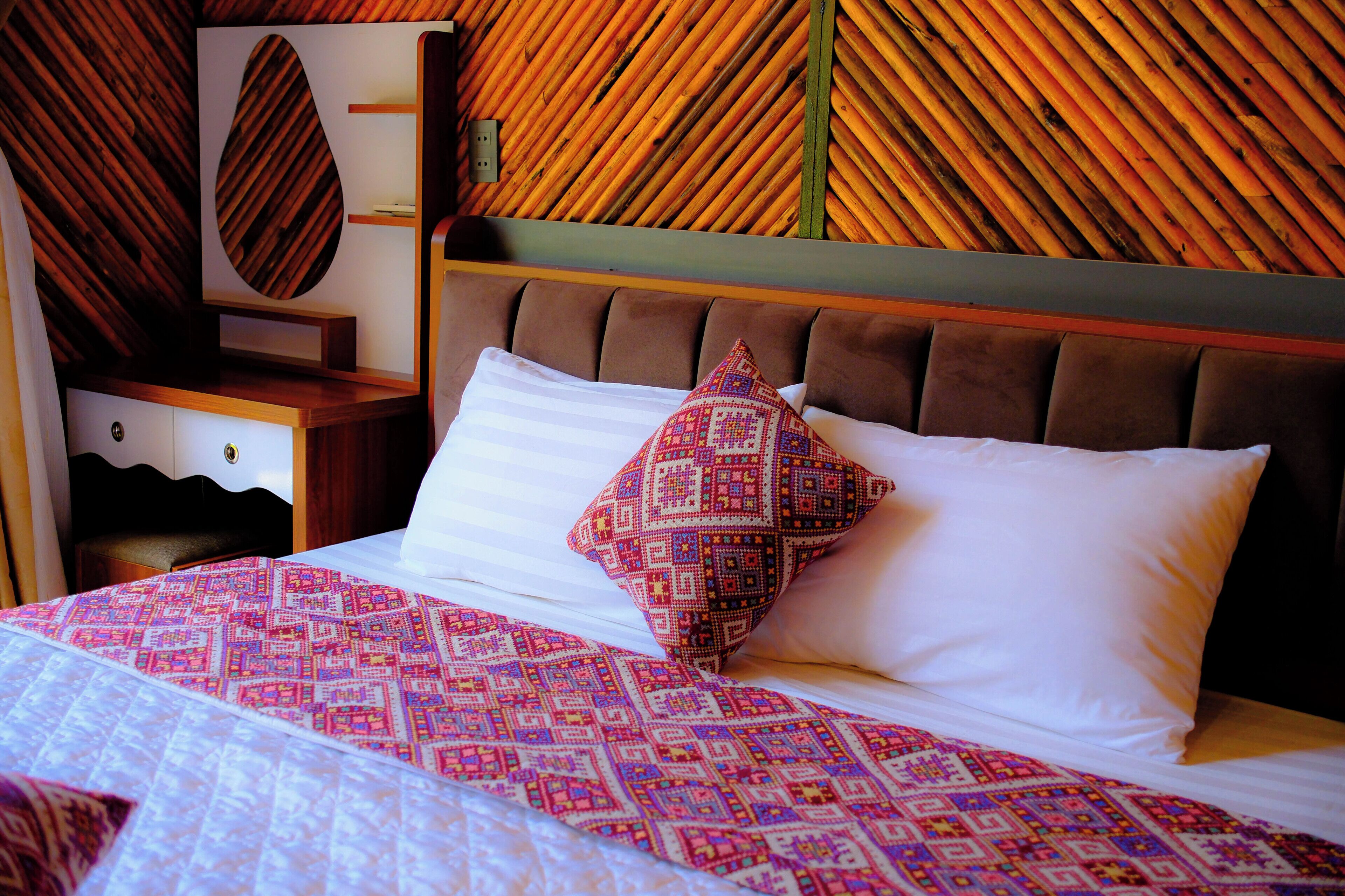A bed with a patterned bedspread, two pillows, a wooden headboard, and a wall with a bamboo-like pattern.