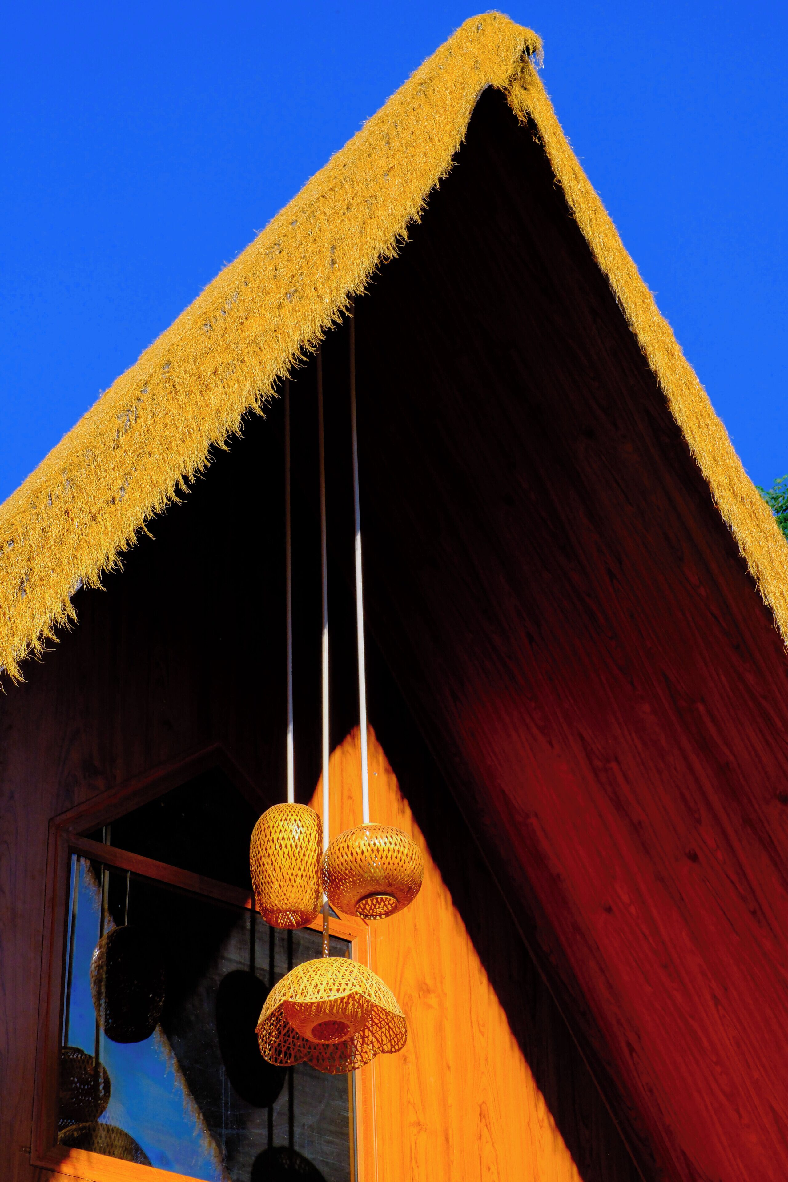 A wooden cabin with a thatched roof, hanging woven light fixtures, and a window reflecting the sky.