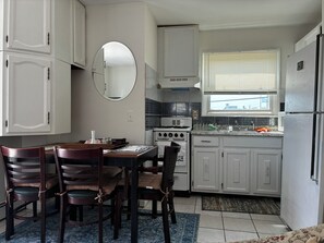 A kitchen with white cabinets, a stove, a sink, and a dining area with chairs and a table. - Lavallette 80s beach nostalgia (Lavallette)