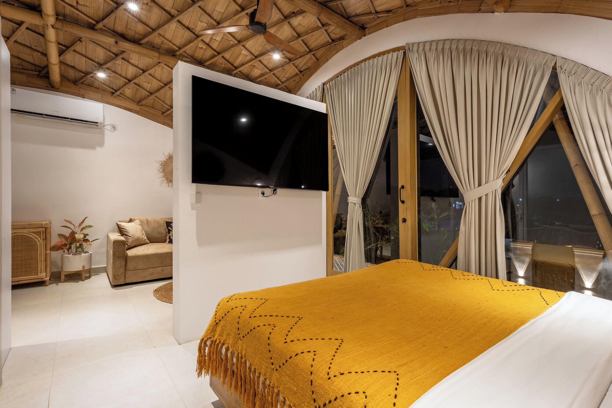 A bedroom with a bed, a TV mounted on the wall, a window with curtains, a sofa, and a small table with a plant.
