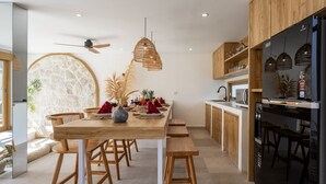 A modern kitchen with wooden cabinets, a central island with seating, and a dining area with a stone wall and a ceiling fan. - New Villa in Kerobokan - 5 Min to Canggu (Badung)