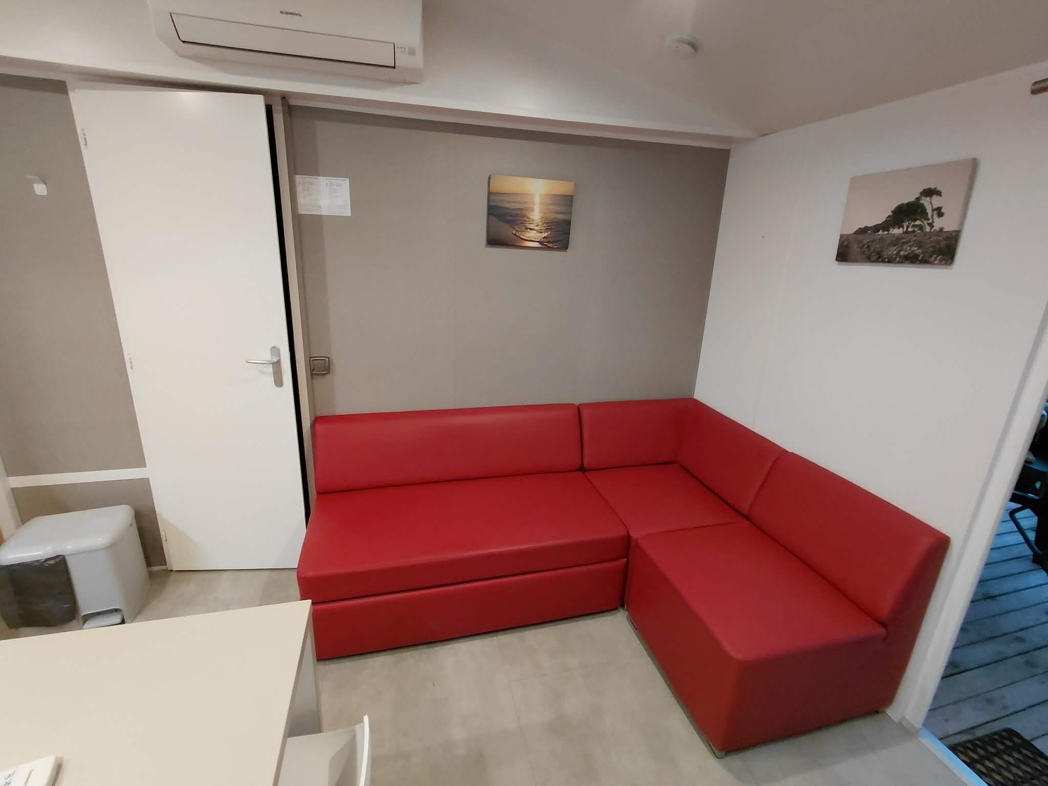 Een rode modulaire bank in een kamer met een witte deur, een klein wit tafeltje en een wandairconditioner.