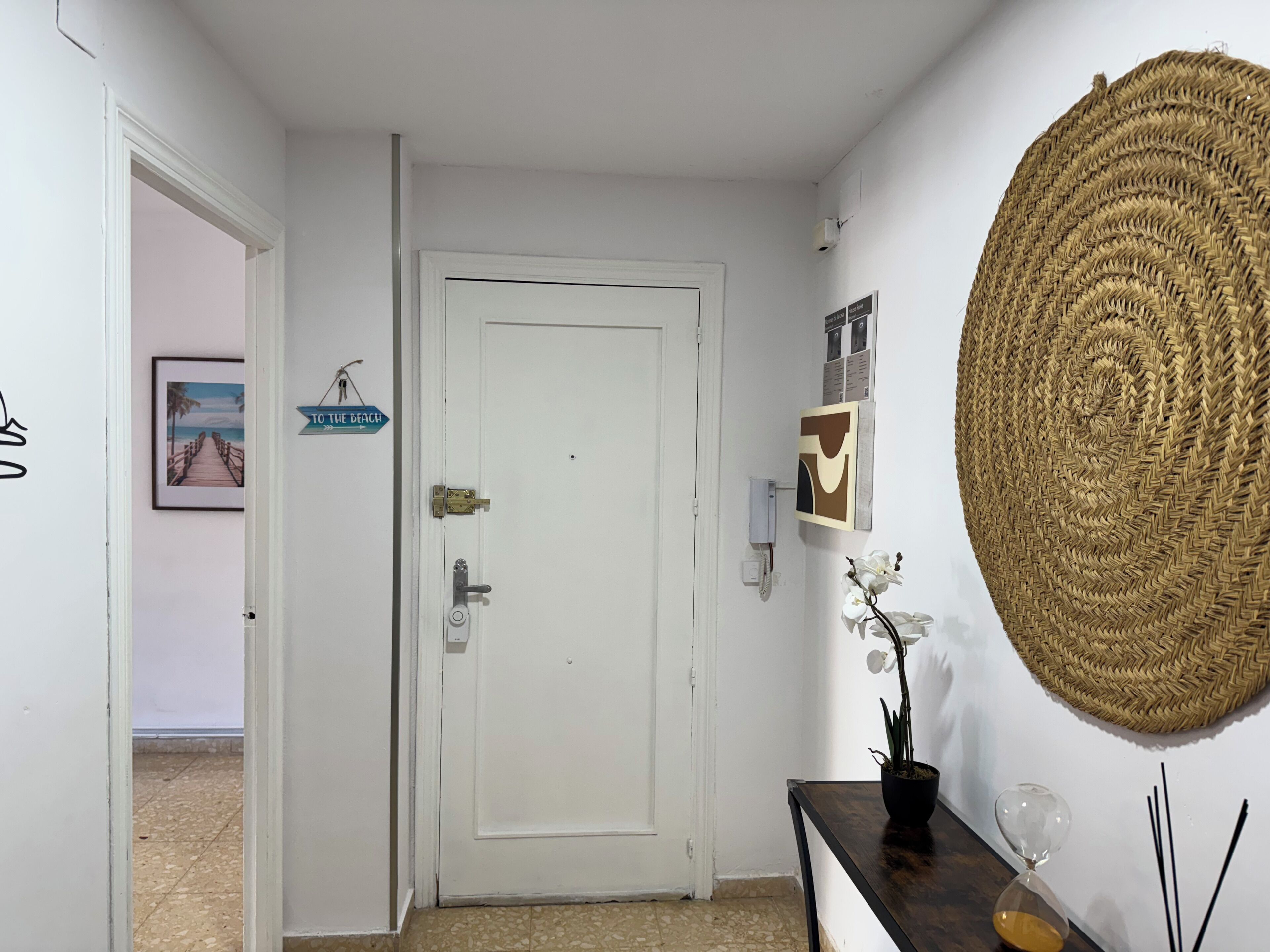 Un pasillo con una puerta blanca, un cuadro enmarcado, un cartel, un tapiz de pared tejido, una orquídea en maceta y una esfera de cristal con arena sobre una mesa de madera.