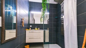 Uma casa de banho moderna com azulejos escuros, cabine de duche em vidro, lavatório branco e espelho.